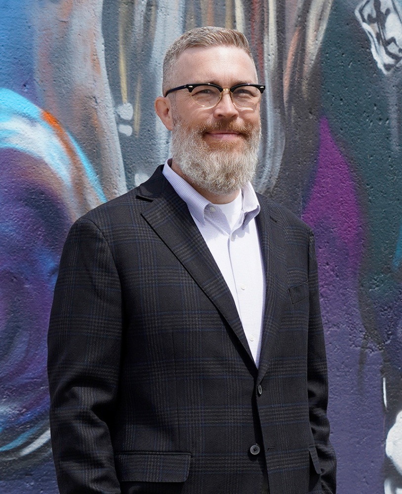A man wearing a dress shirt and a sport coat poses as he looks at camera while standing in front of a mural on a cement wall.