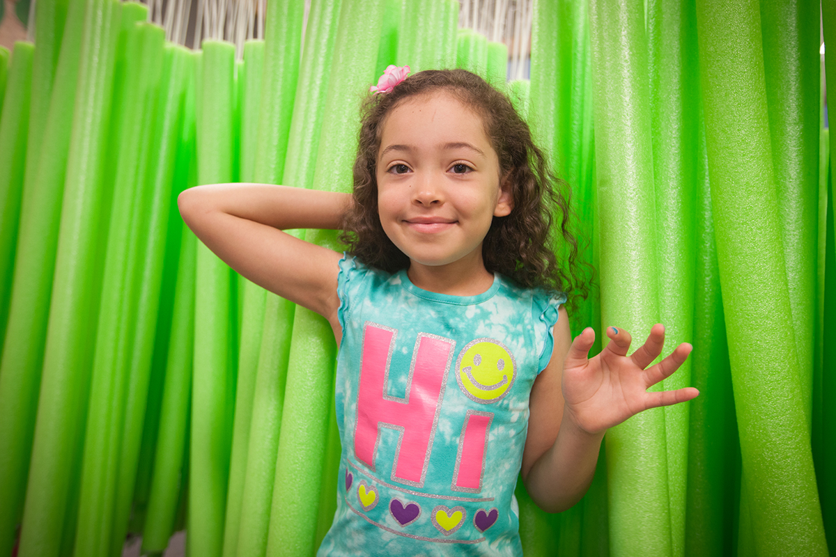 A girl stands in front of a wall of bright green pool noodles at the Flint Farmers' Market.