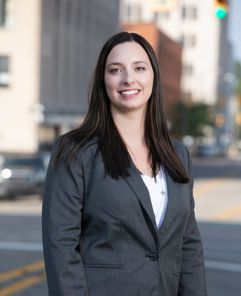 A young woman who wears a grey suit jacket and white top poses for the camera outdoors.