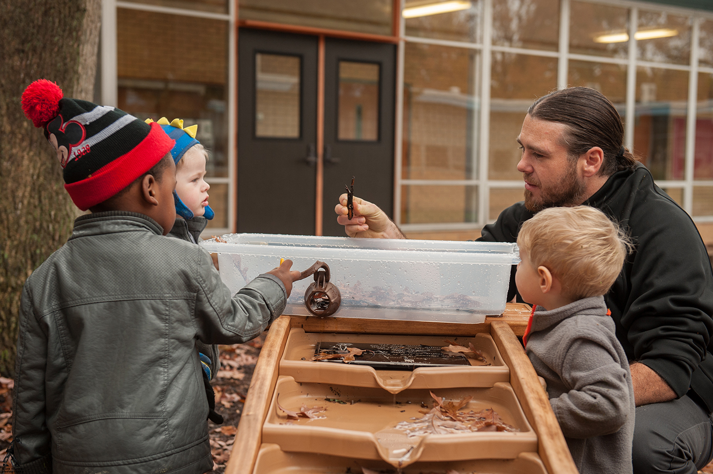 A group of children play at an outdoor water table while a man shows them a small leaf.