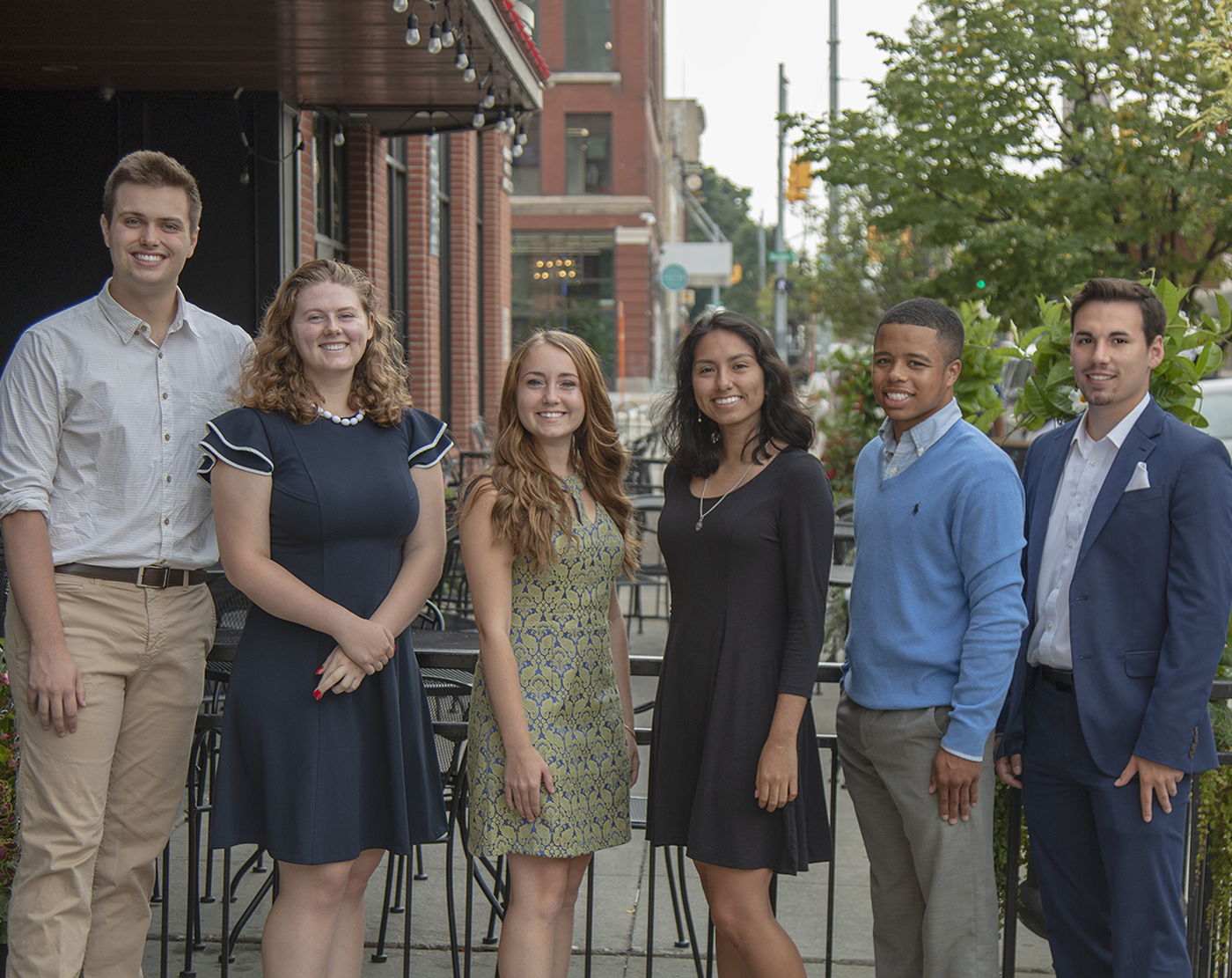 A smiling cohort of 2018 interns stands in a row in front of a row of brick buildings.