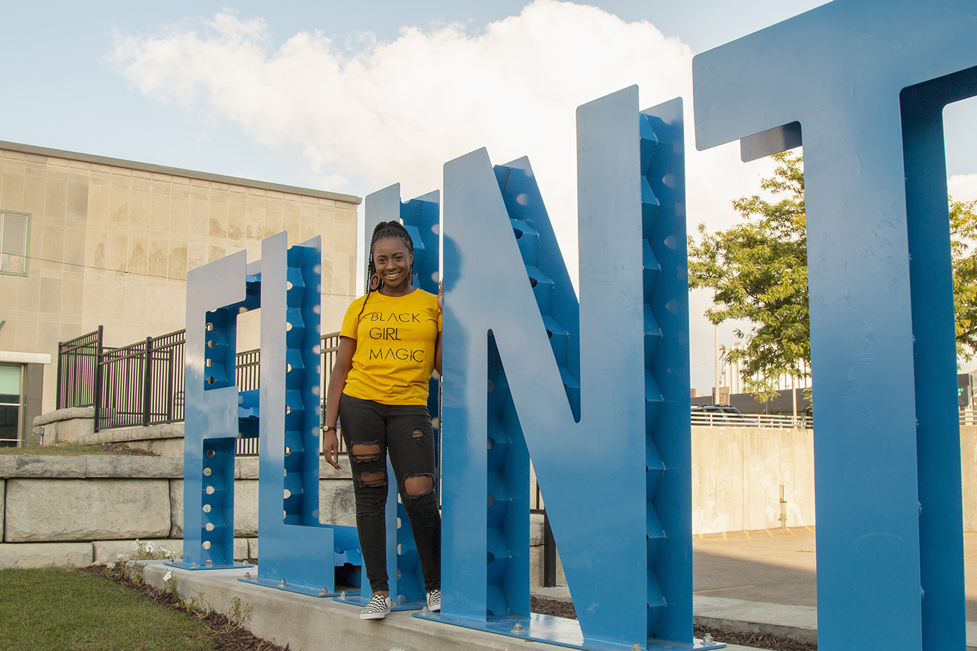 Artist and poet Kayla Shannon stands smiling in front of large blue letters spelling out Flint.
