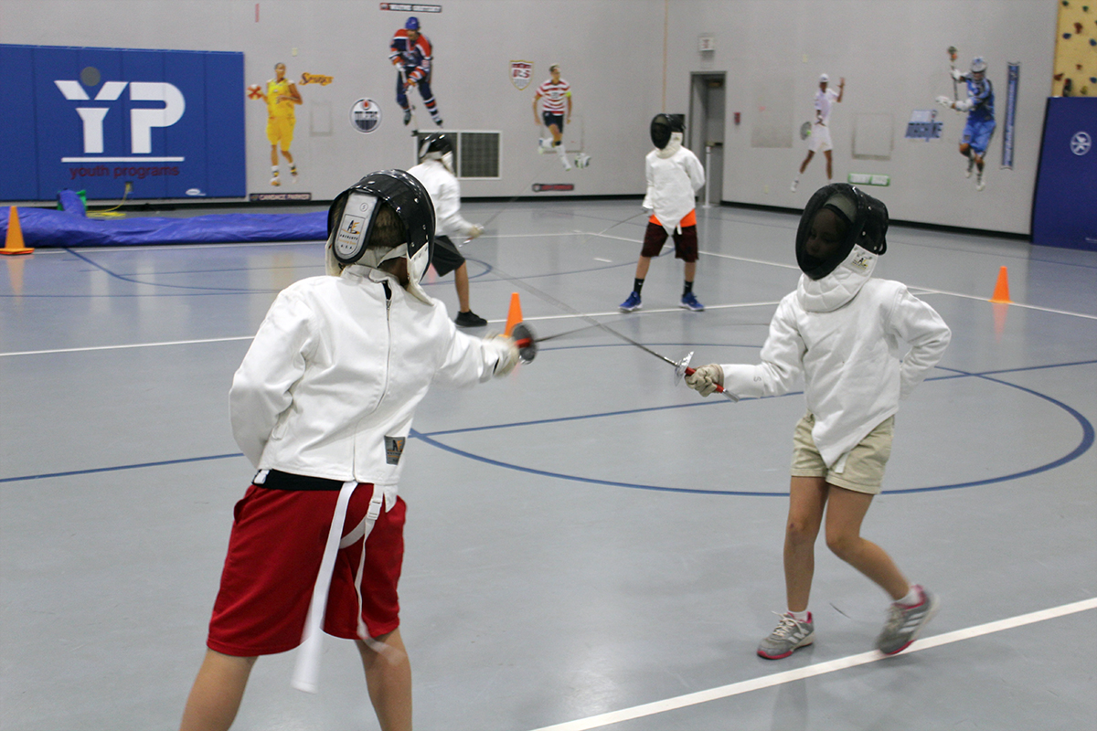 Two young students standing in a gym wearing full fencing gear clash swords. 