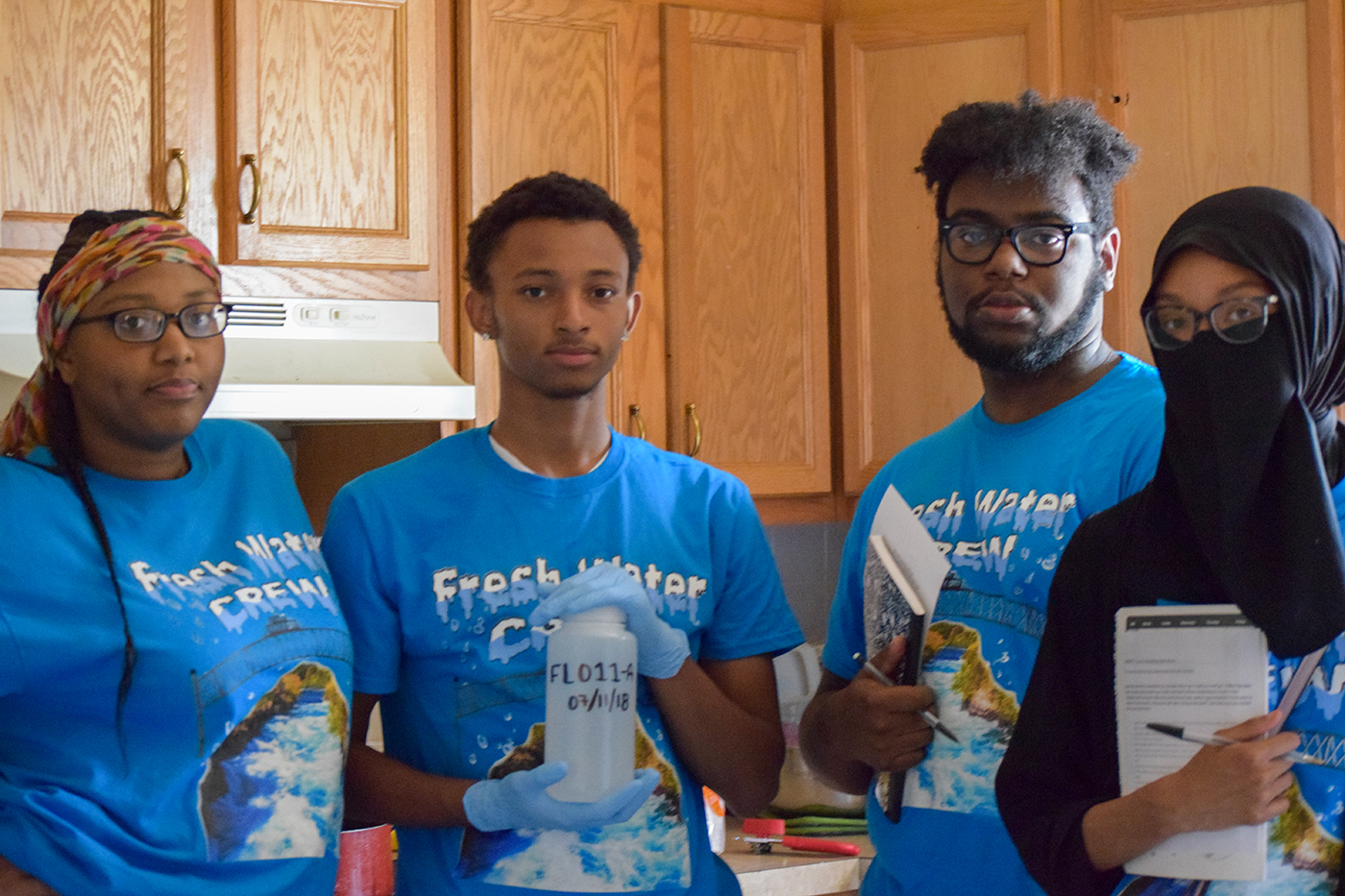 A group of four young people wearing matching Fresh Water Crew tshirts stand in a kitchen holding input forms and a water testing bottle.