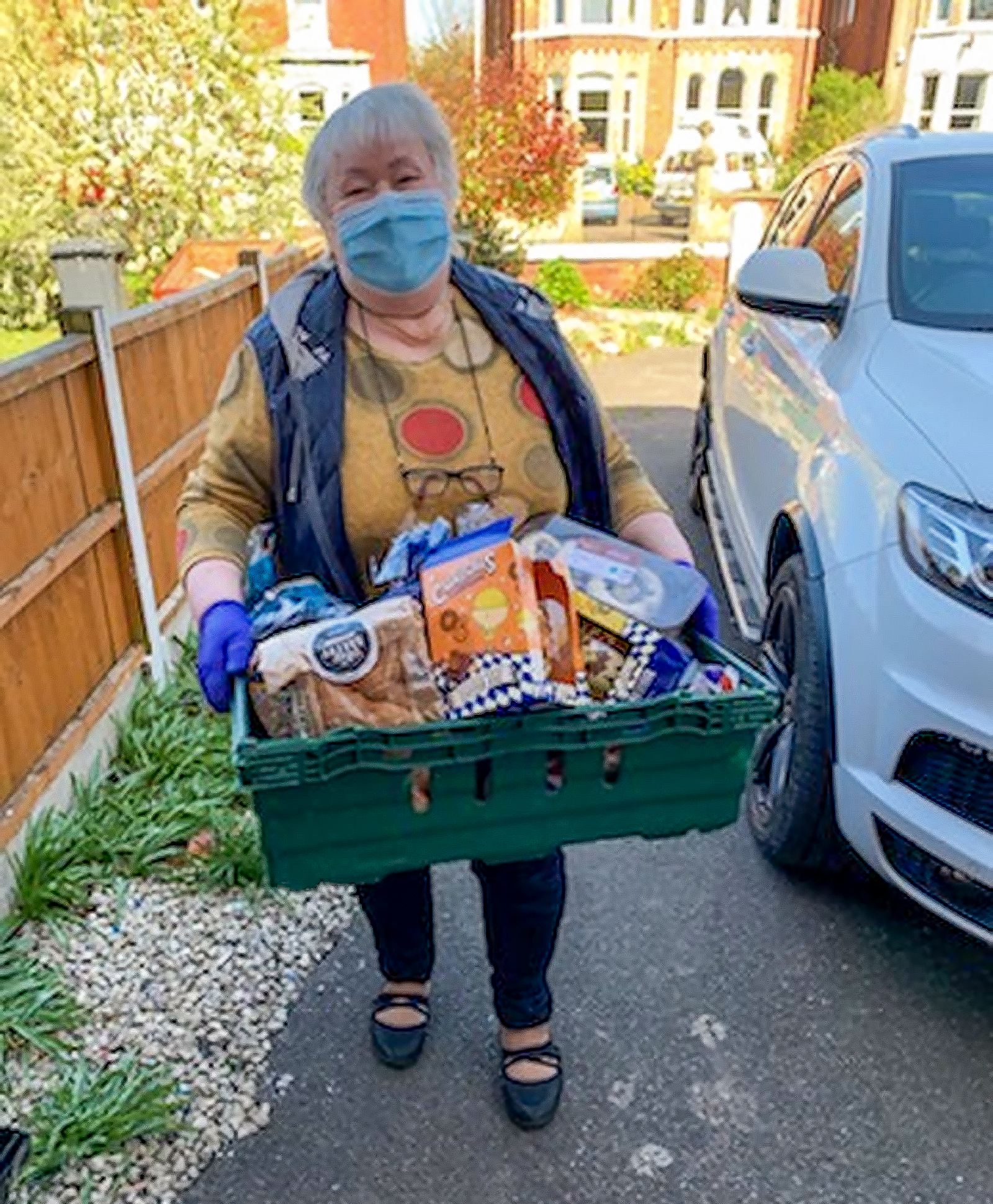 A volunteer that is wearing a facemask and a colorful sweater stands in a driveway while holding a box of goods that is ready to be delivered to people affected by the coronavirus shutdowns.