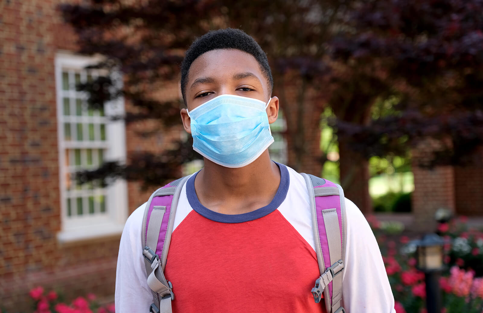 An African American teenager wears a backpack and facial mask to protect himself from COVID-19. He is standing in front of a brick building and pink flowers.
