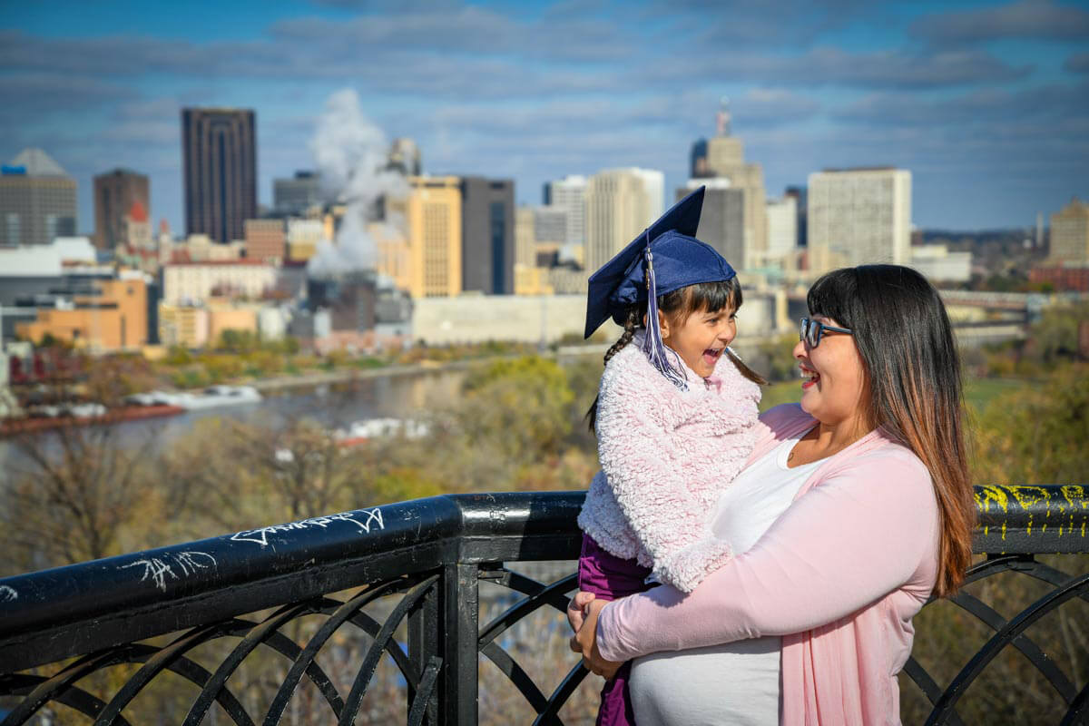 A parent and her child stand in front of a city skyline. The child is smiling and wearing a graduation cap.