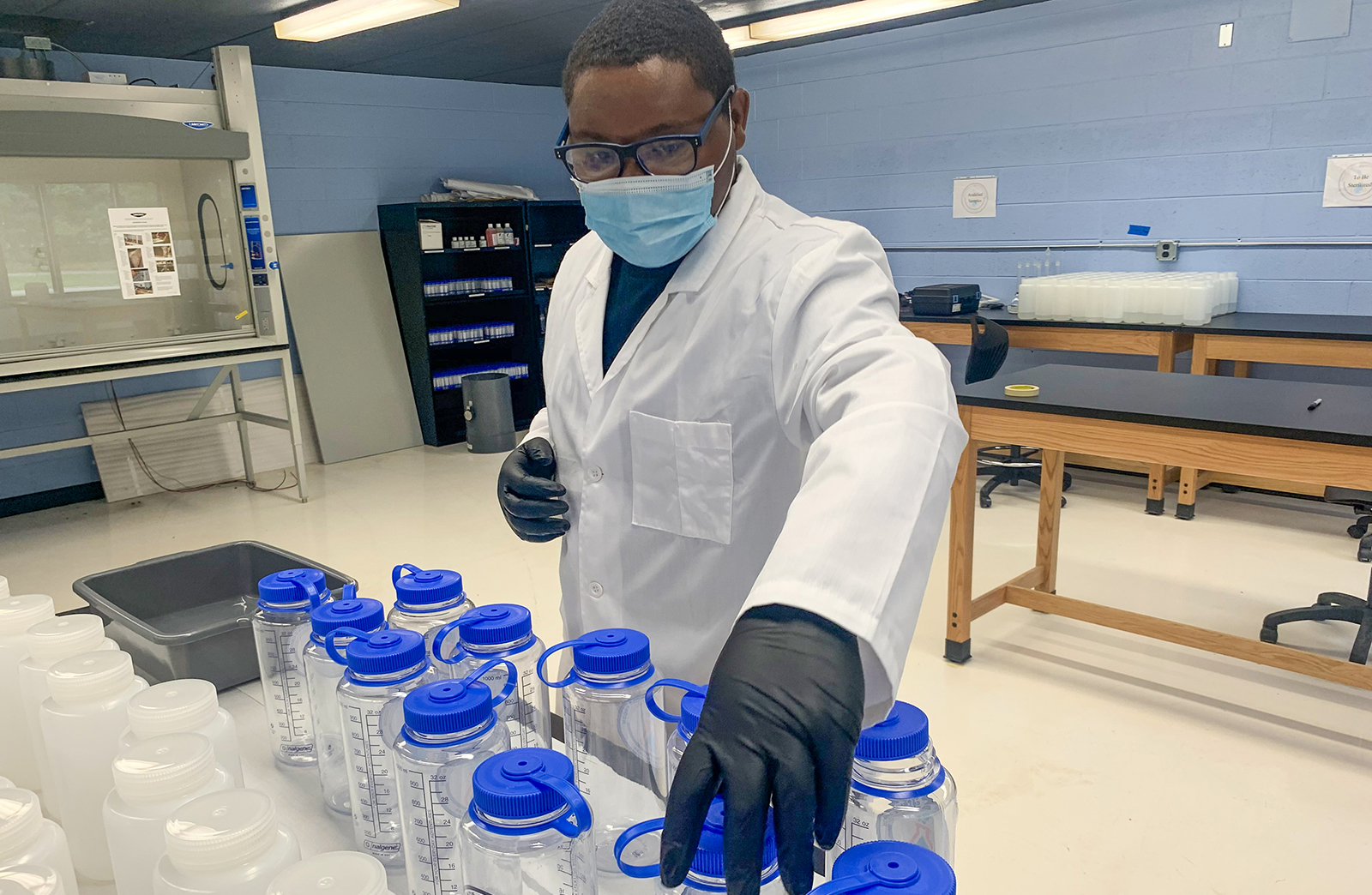 A student wearing a mask, white lab coat and gloves works with water testing bottles in the lab at the McKenzie Patrice Croom Memorial Flint Community Lab.