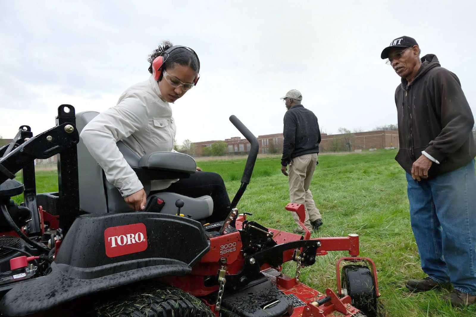 A young woman is seated on a lawnmower. Two men stand nearby.