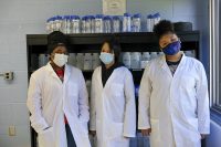Three young women stand in front of a lab shelf that holds labeled water samples.