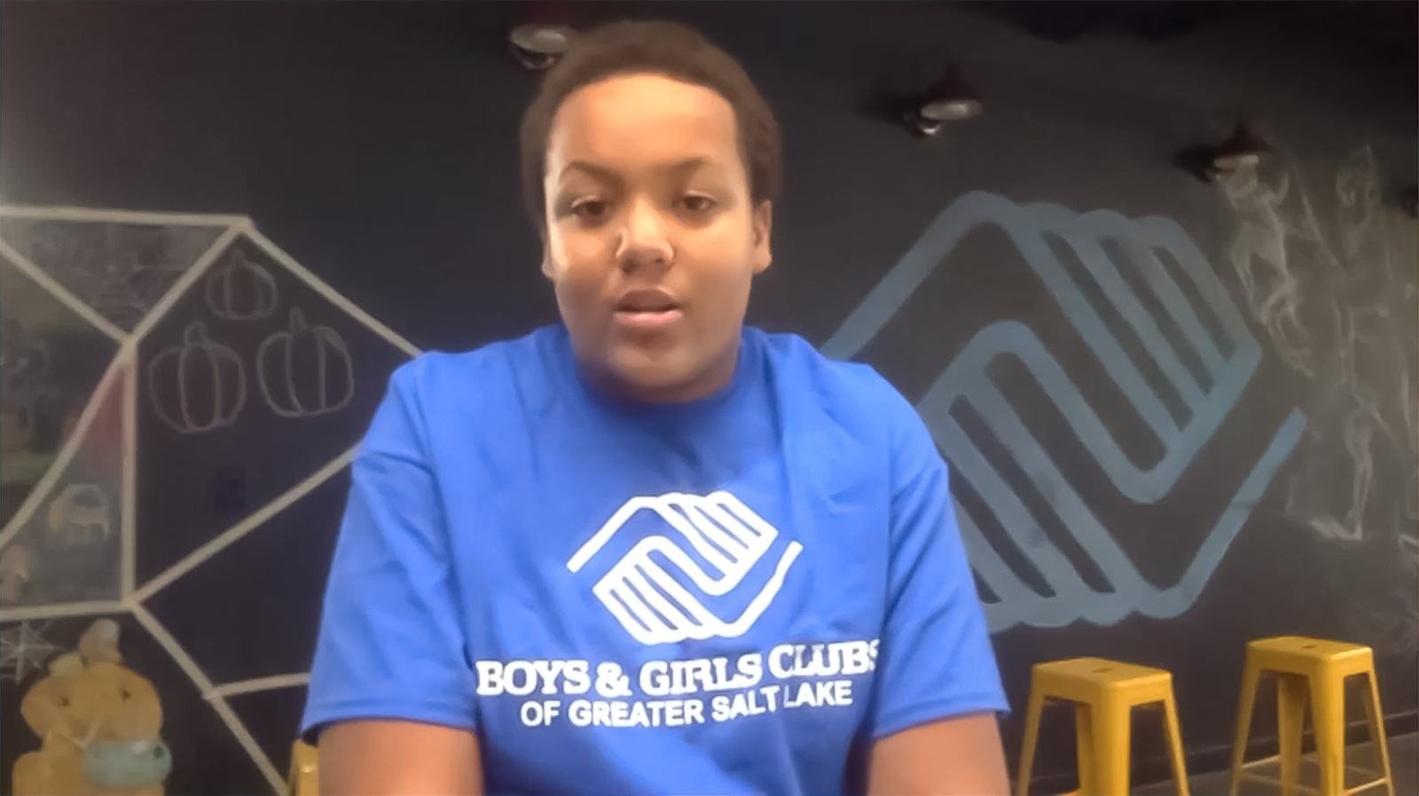 A boy wearing a Boys & Girls Club tshirt sits in front of a chalkboard.