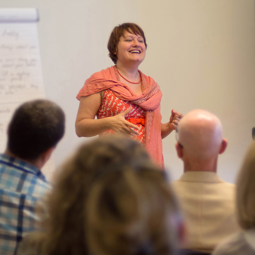 A smiling woman gives a presentation to a room of seated people.