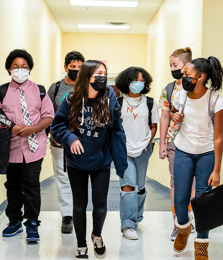 A group of students walk down a hall. They all wear masks.