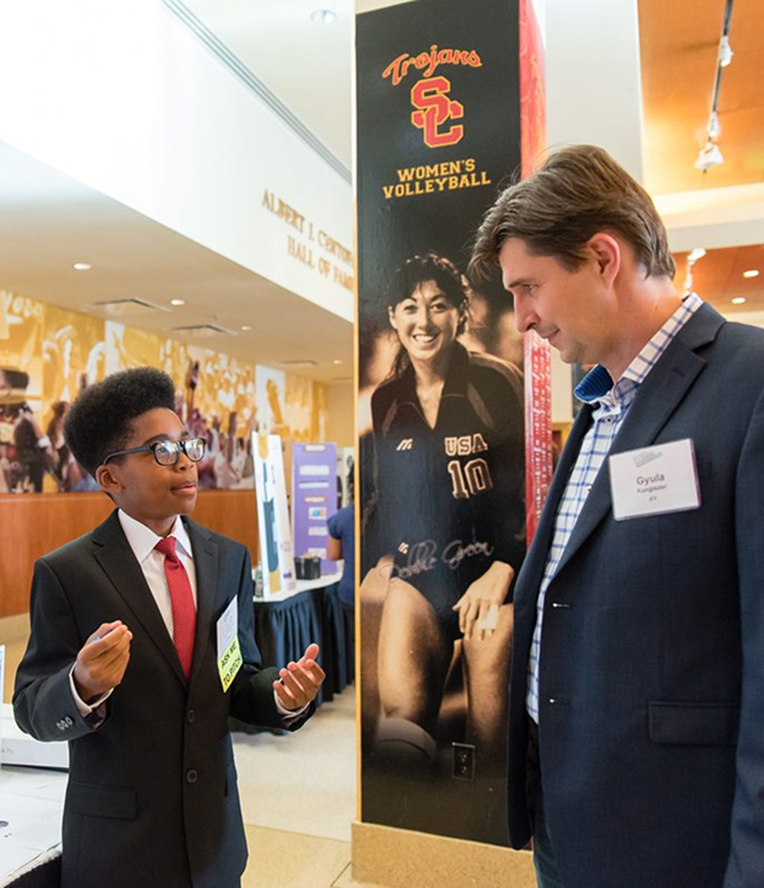 A boy wearing a suit and standing in front of a tabletop presentation makes an entrepreneurship pitch to a man as part of a regional youth challenge.