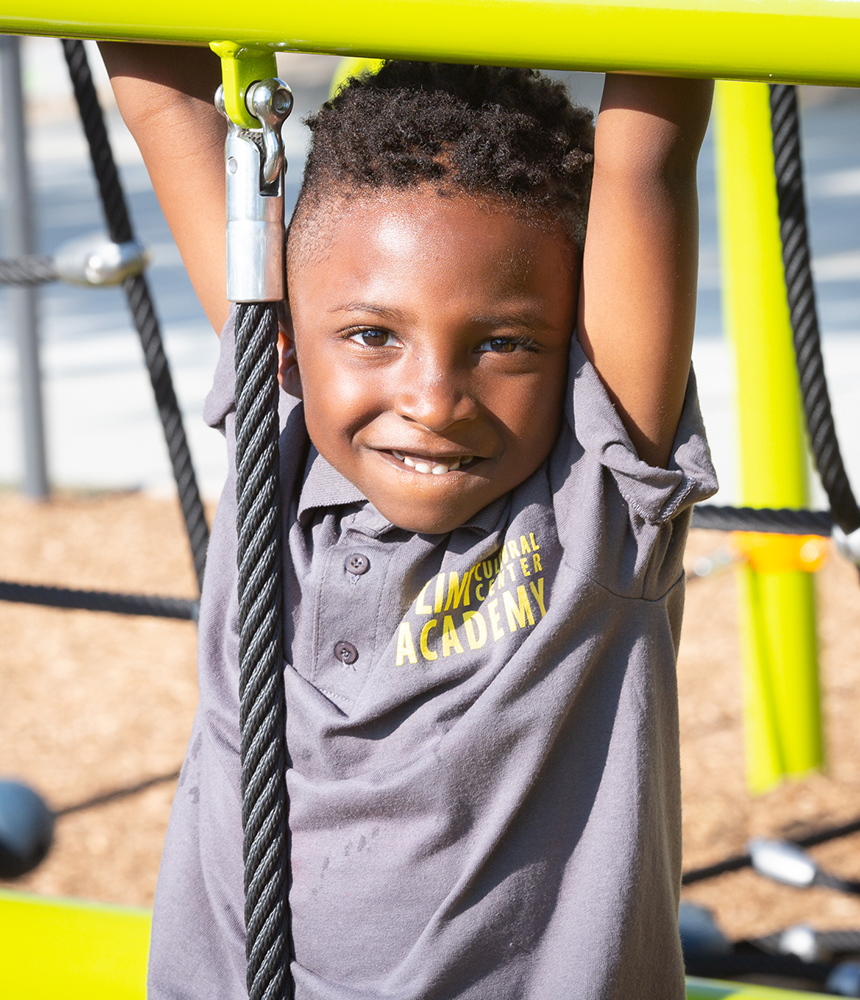 A smiling young boy hands from an outdoor gymnasium. He wears a shirt with the text Flint Cultural Center Academy.