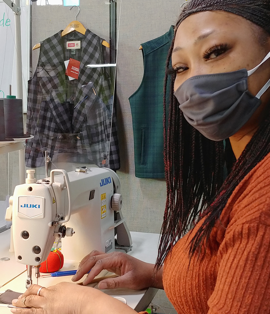A woman wearing a mask sits at a sewing machine. She looks at the camera. Two vests hang on the wall behind her.
