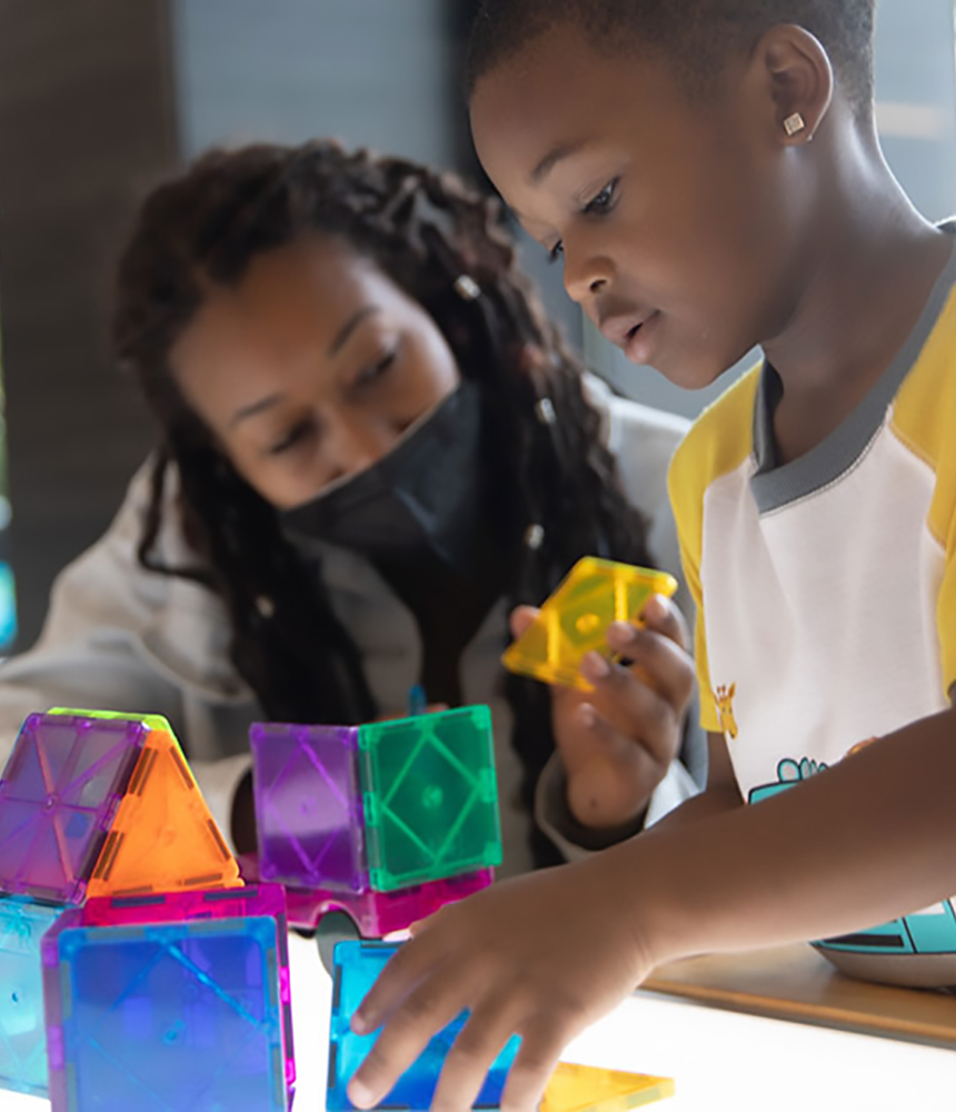 A young Black child is playing with lit up blocks on a table in the Flint Public Library's children's section while an adult woman wearin a mask looks with interest at his progress.