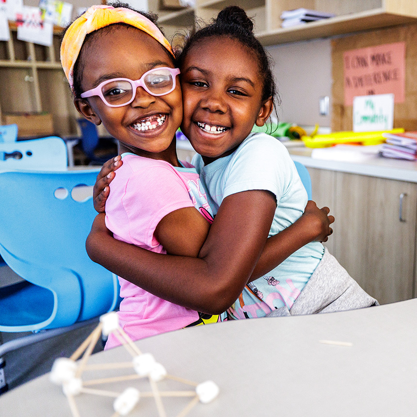 Two girls are hugging happily while sitting in a bright classroom, surrounded by colorful seats and craft supplies. Two small-scale models of molecules or atoms constructed using toothpicks and small marshmallows are placed in front of them on a light grey table.
