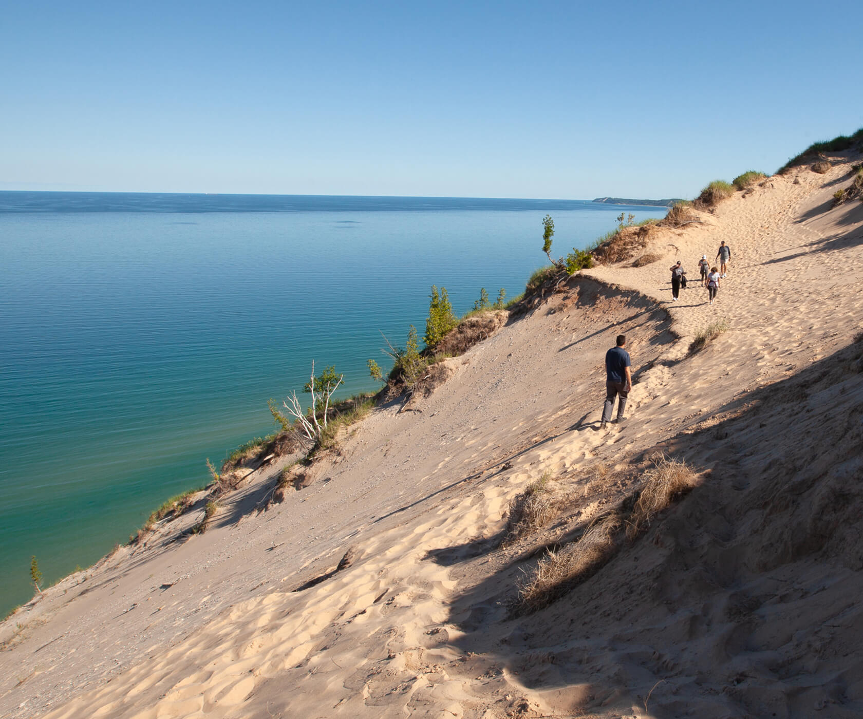 A group of people walk along a scenic, large sandy dune that runs along a shoreline.
