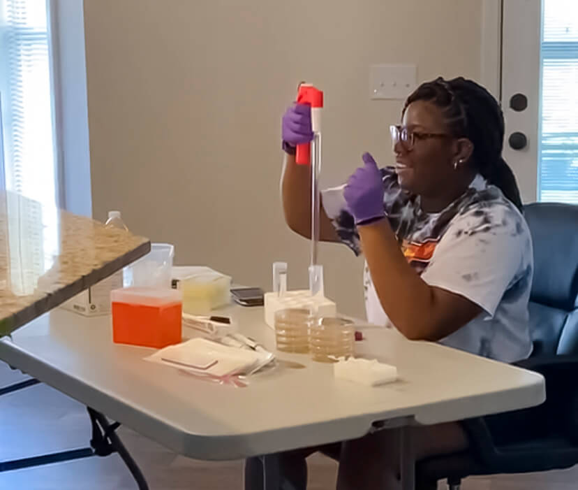A college aged Black female student pipettes a substance into a test tube at a makeshift laboratory set up in her home's living room for her internship during the COVID-19 pandemic.