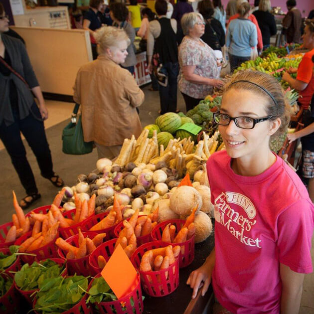 A young woman looks up and smiles while working at a produce booth at the Flint Farmers' Market.