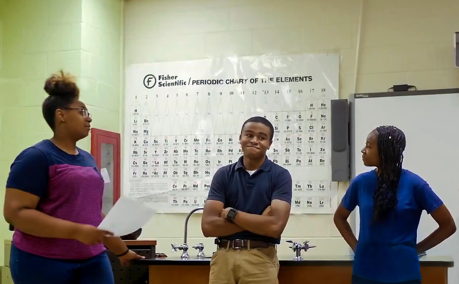 Three Black students hone their presentation skills at the front of a lab room that has the periodic table hanging above a laboratory counter located behind them.