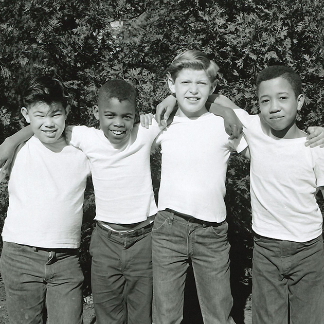 An old black and white photo shows four smiling young boys in white tshirts standing arm in arm.