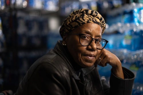 A side profile portrait of a mature Black woman wearing a head wrap and glasses looks at the camera.