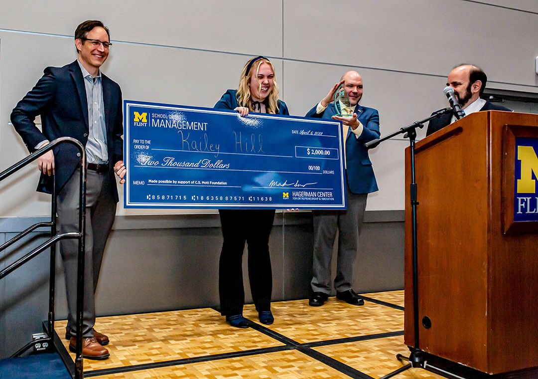 A woman holds a giant check made out for $2,000 from the UM-Flint School of Management. Three men join her on the stage. One holds a glass trophy and one stands behind a podium.