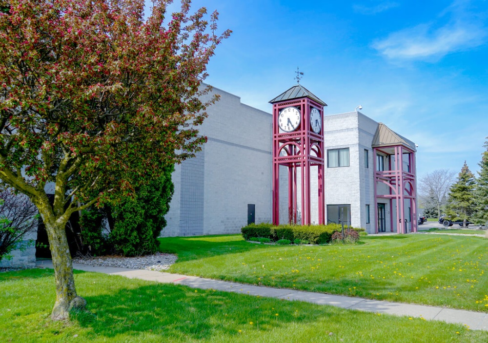 An office building sits in a grass-filled lot dotted with dandelions. A large ornamental red clock with a weathervane on the top is in front of the building.