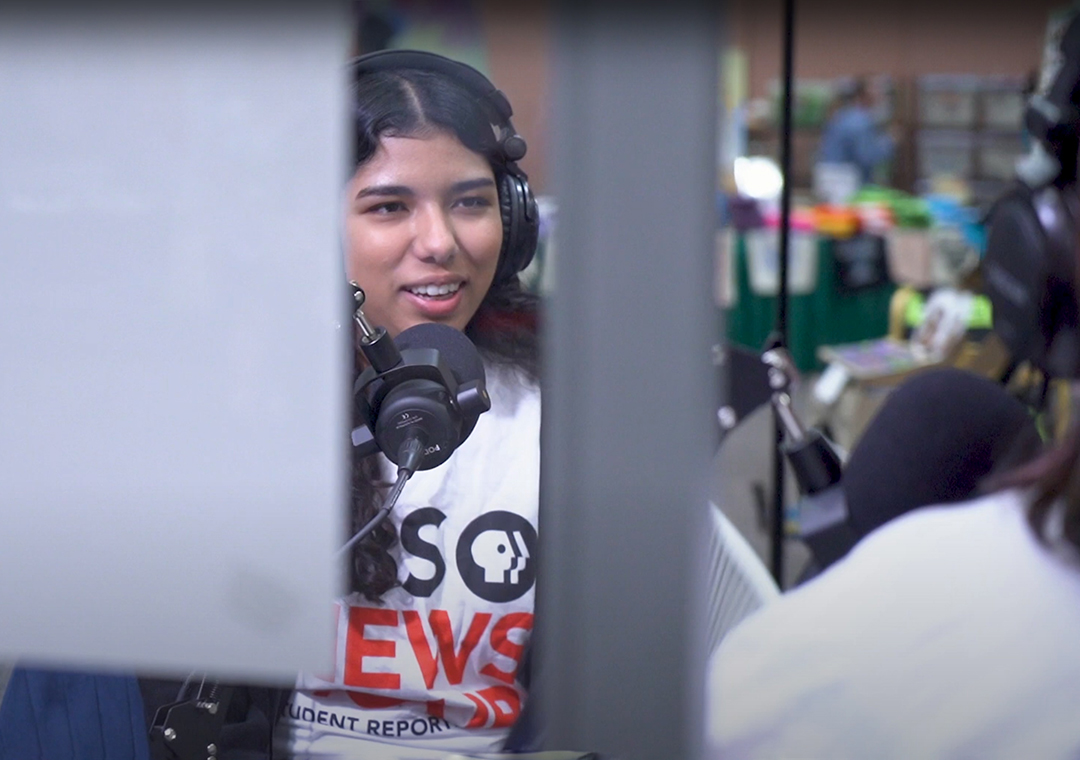 A student reporter wearing a PBS NewsHour tshirt sits at a microphone while talking with another reporter in a recording lab.