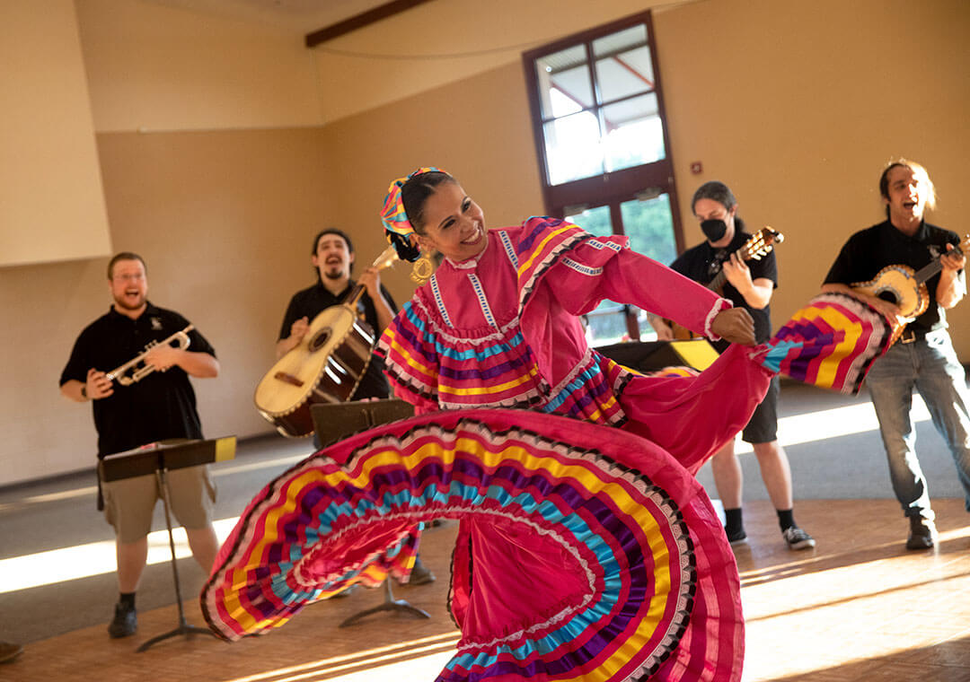 A traditional folkloric performer in a brightly colored dress covered in ribbon strips happily dances to the music of the mariachi playing behind her.