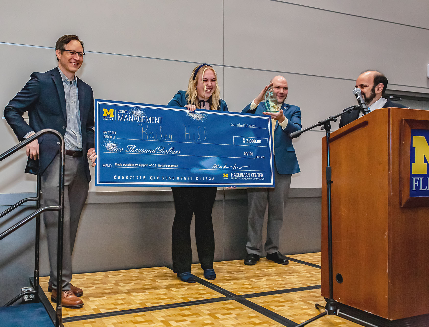 A high school student, Kailey Hill, stands on a raised stage holding after having been handed by officials from University of Michigan-Flint’s Zillion Solutions program a large carboard check made out to her for $2000.