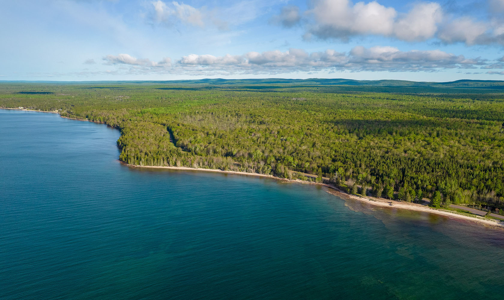 An aerial view of the Lake Superior shoreline with crystal clear deep blue/green water showing the underwater fauna at Little Betsy Point and the neighboring forest.