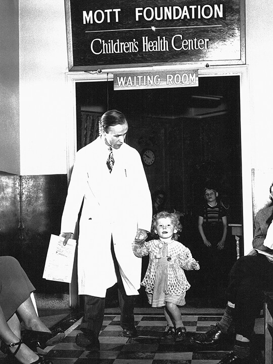 A 1930s era black and white photo shows a doctor walking a small girl through the doorway under a sign that reads Mott Foundation Children's Health center Waiting Room.