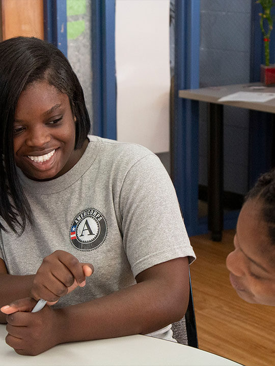 AmeriCorps member Jani Toney smiles at a young student. The two are sitting at a table together.