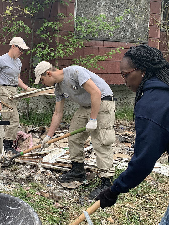 AmeriCorps members use shovels to clear away debris from the yard of an abandoned home.