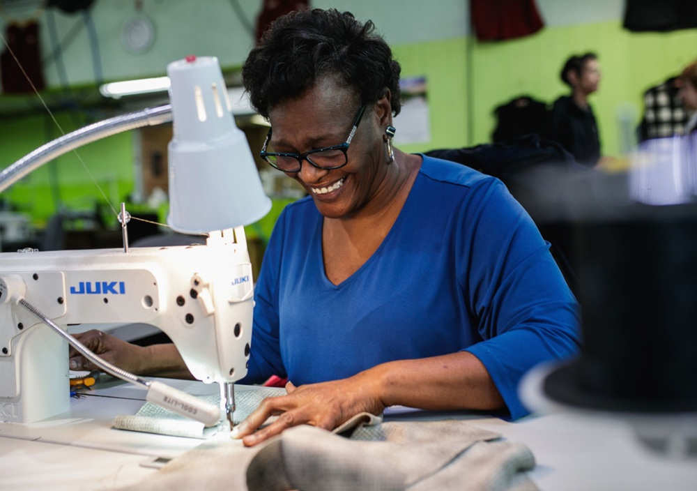 A woman sews a vest at a Juki professional sewing machine while smiling down at her work.