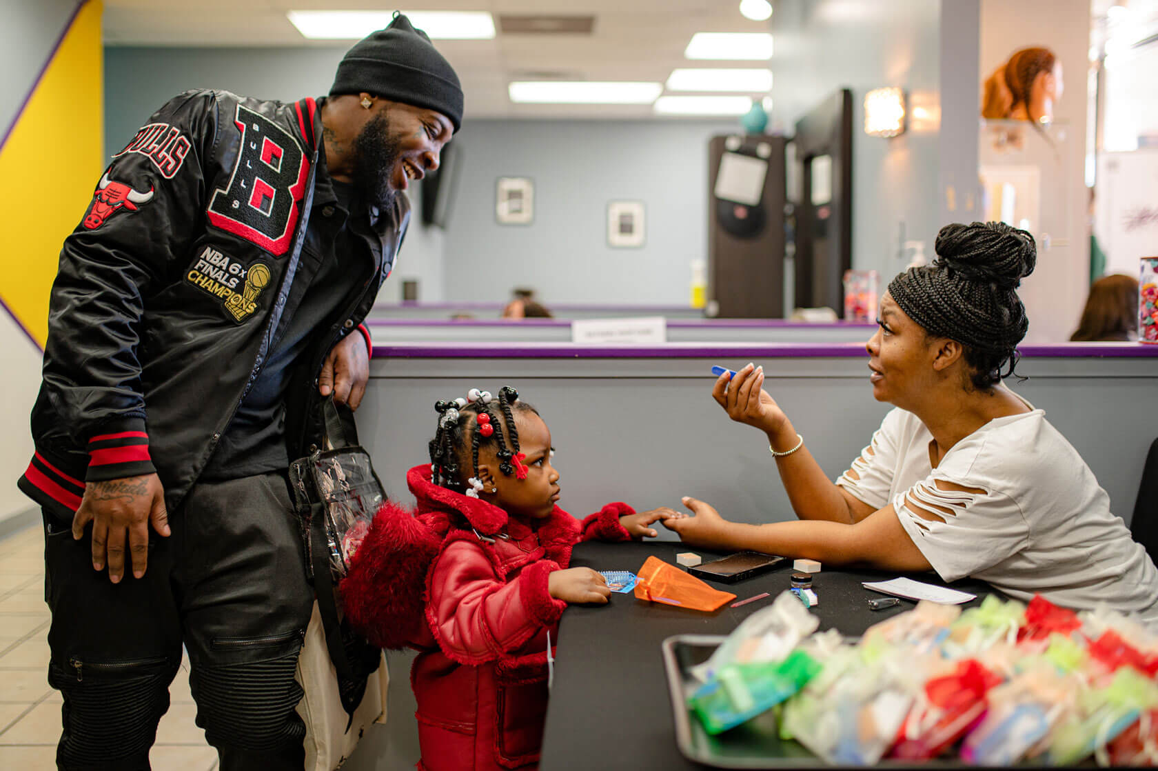 A smiling and engaged man watches a woman use an emery board on the fingernails of his very young daughter.