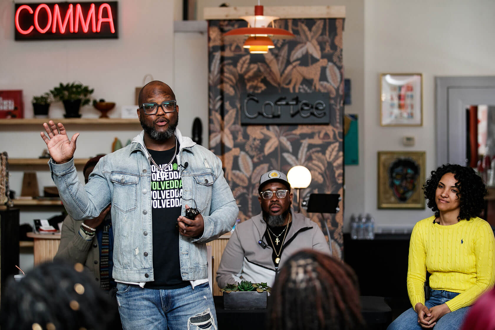 A standing man raises his right hand while speaking to a group in a comfortable looking coffee shop. A man and a woman are seated behind him.