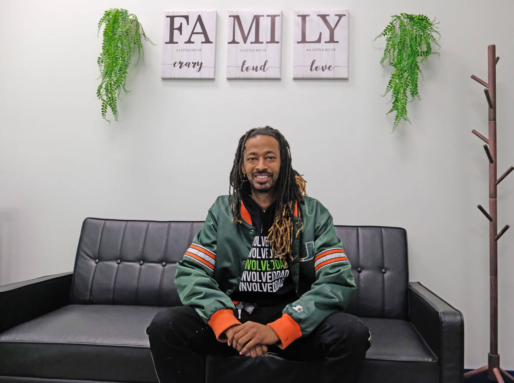 A smiling man wearing an "InvolvedDad" t-shirt sits on a couch below a set of signs reading "Family, a little bit of crazy, a little bit of loud, a little bit of love."