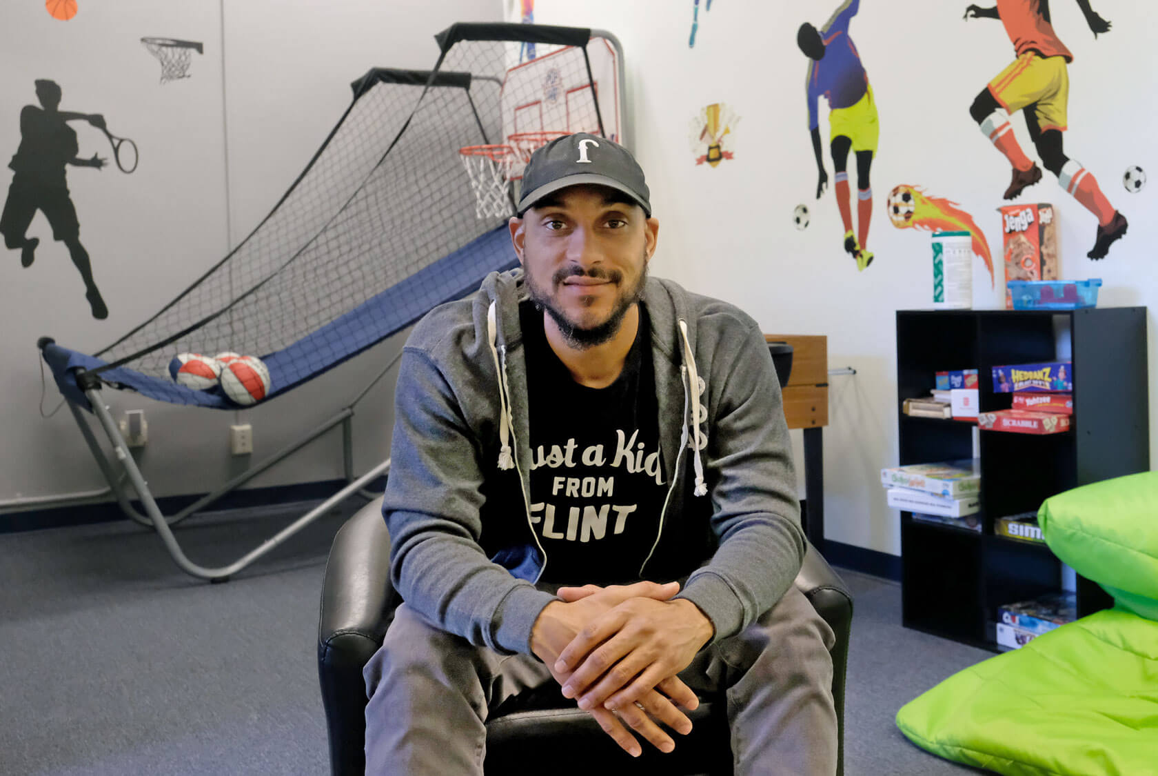 A young man with a small smile wearing a shirt that reads "Just a kid from Flint" sits in a recreation room. Murals of figures playing sports decorate the walls. A basketball game and shelf with board games are in the background, along with bright green cushions.