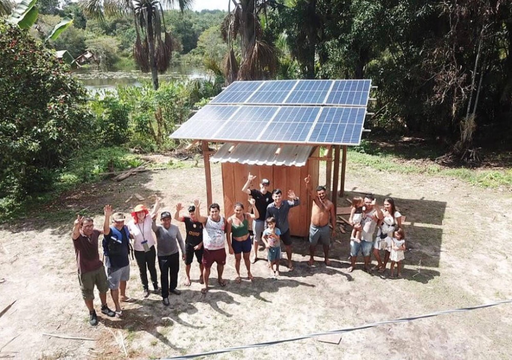 An aerial image of eight solar panels installed on stilts with a small utility housing unit below it. There is a group of 12 adults and three children waving at the drone's camera as it hangs in the air and takes their photo. The backdrop is the tropical forest with a river running through it.