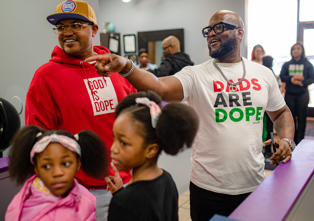 A smiling man wearing a "Dads are Dope" t-shirt points out something outside the frame to a man and two girls.