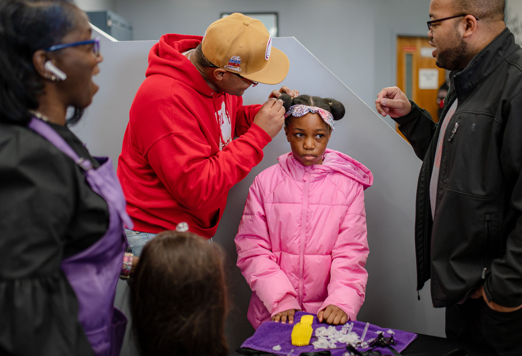 A girl wearing a bright pink coat stands patiently while her dad works on her hair. A woman and man watch from the sides.