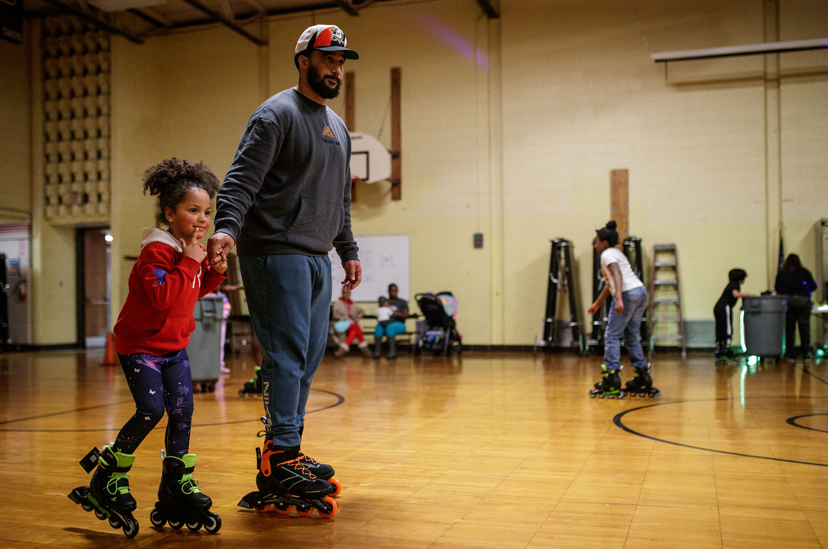 A dad and his daughter rollersjate in a school gym.