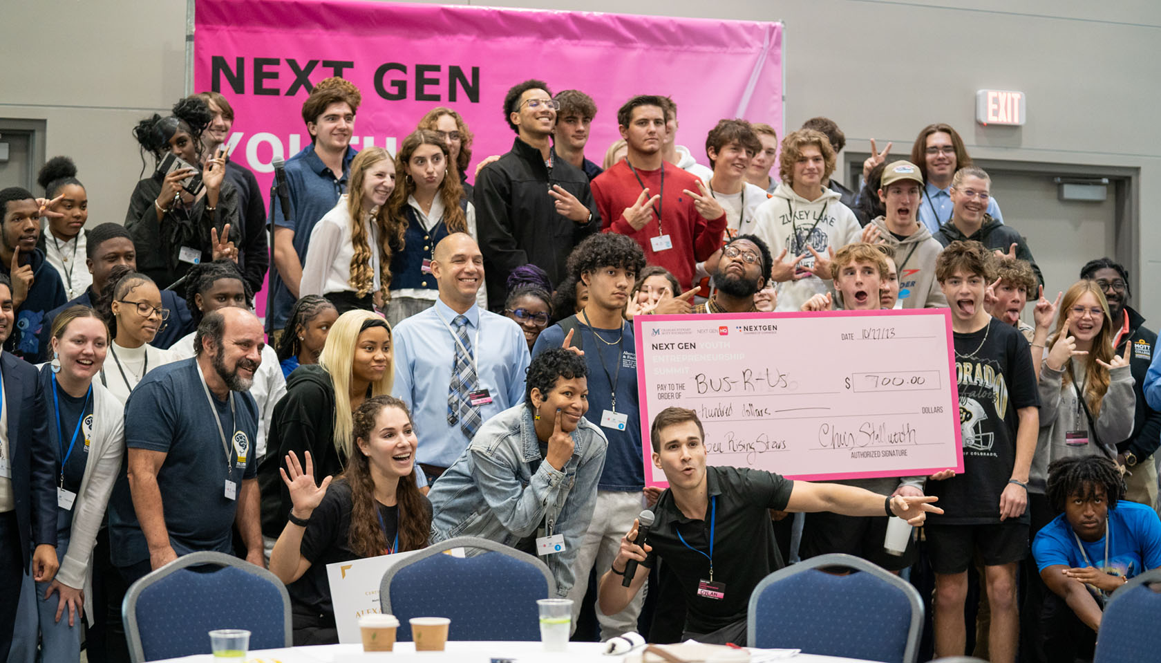 A group of young people that attended the NextGen Youth Entrepreneurship Summit event and their mentors pose on a stage for a congratulatory group shot.