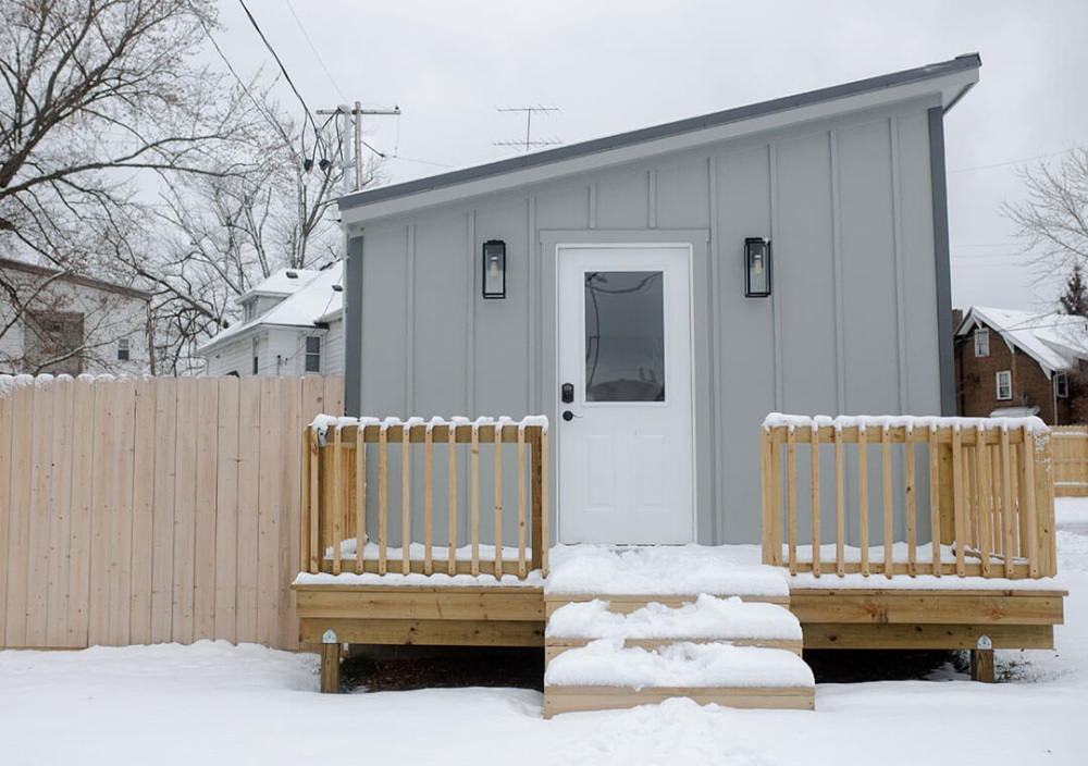 A small gray home with a white door and a wood porch and fence, photographed on a snowy, gray day.