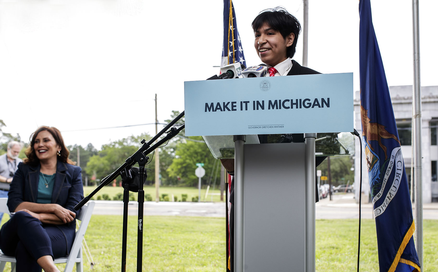 A smiling young man speaks into several microphones at a podium with a Make it in Michigan sign attached to the front. A Michigan flag is next to him, and the Latinx building is in the distance. Gov. Whitmer sits in the background.