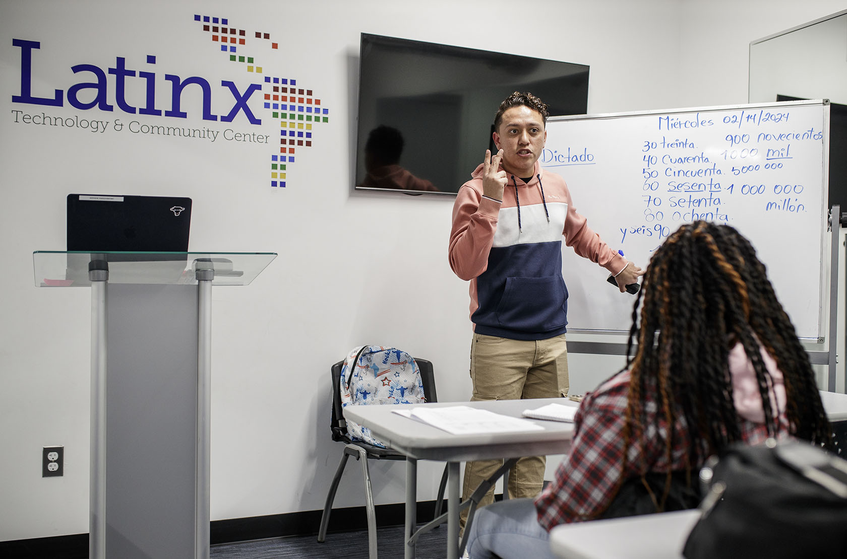 A man holds up two fingers and points to a whiteboard while speaking to a group in a classroom style setting. A Latinx Technology & Community Center logo is on the wall.